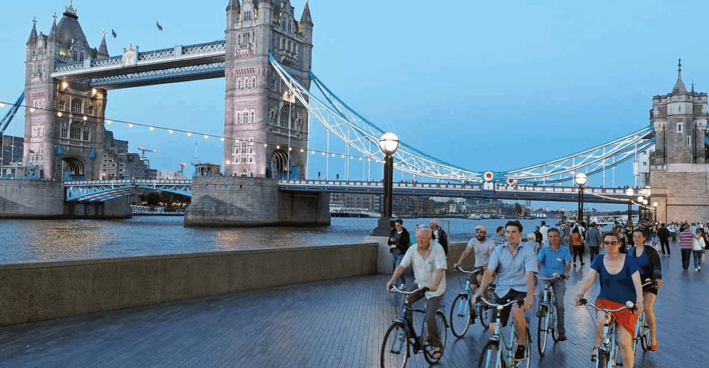 A group adults on bicycles along the banks of the Thames with the Tower Bridge in the background early evening on a London Bike Tour